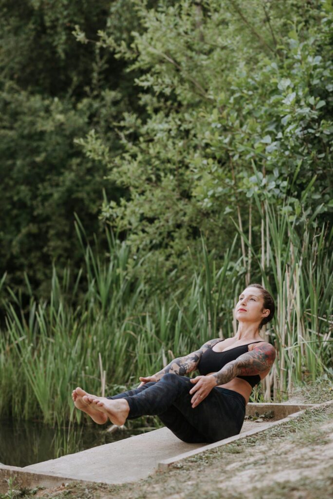 Slim barefoot woman performing Boat pose on a dock.